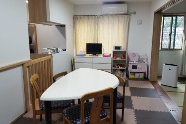 介護施設の共用スペース。テーブル、テレビ、おもちゃ、エアコン完備の明るい部屋。